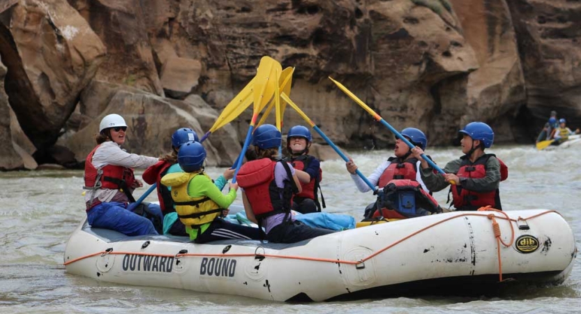 A group of students wearing safety gear sit in a raft and raise their paddles into the air.